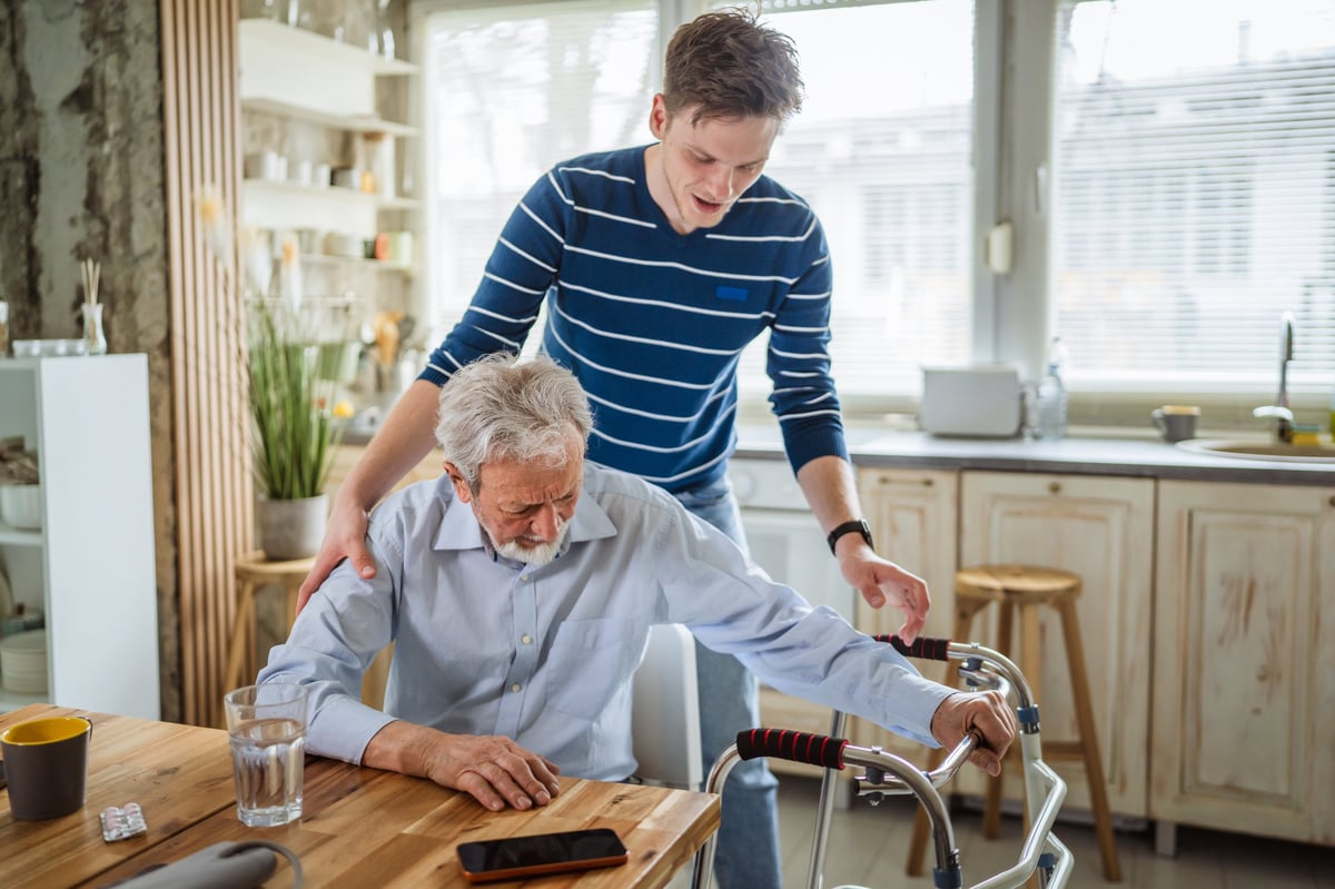 Young man providing support to a senior man with a mobility walker, demonstrating care giving and assistance with daily living in a home kitchen setting
